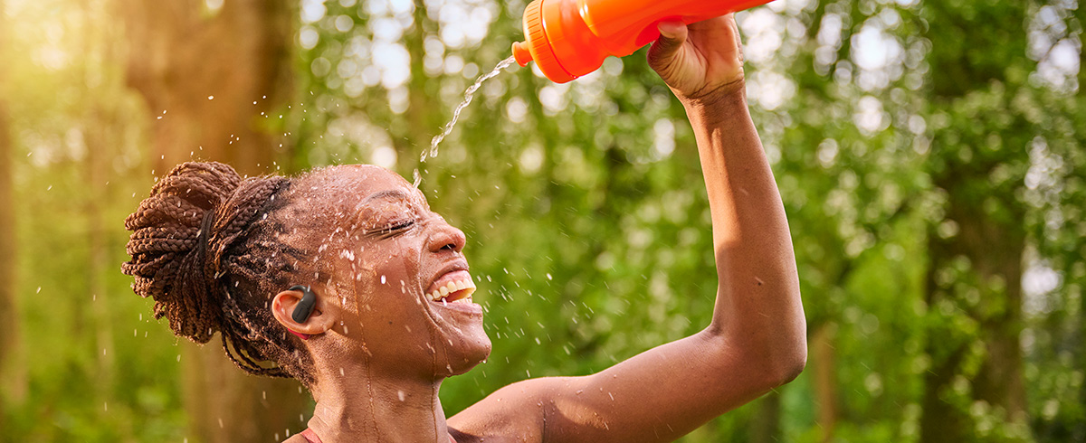 Mujer refresc&aacute;ndose al derramar agua sobre su cabeza mientras usa auriculares deportivos Philips al aire libre