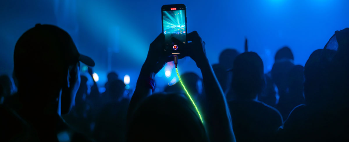 Una persona levanta un tel&eacute;fono m&oacute;vil conectado a un cable brillante frente a una gran multitud, captando el momento durante un evento
